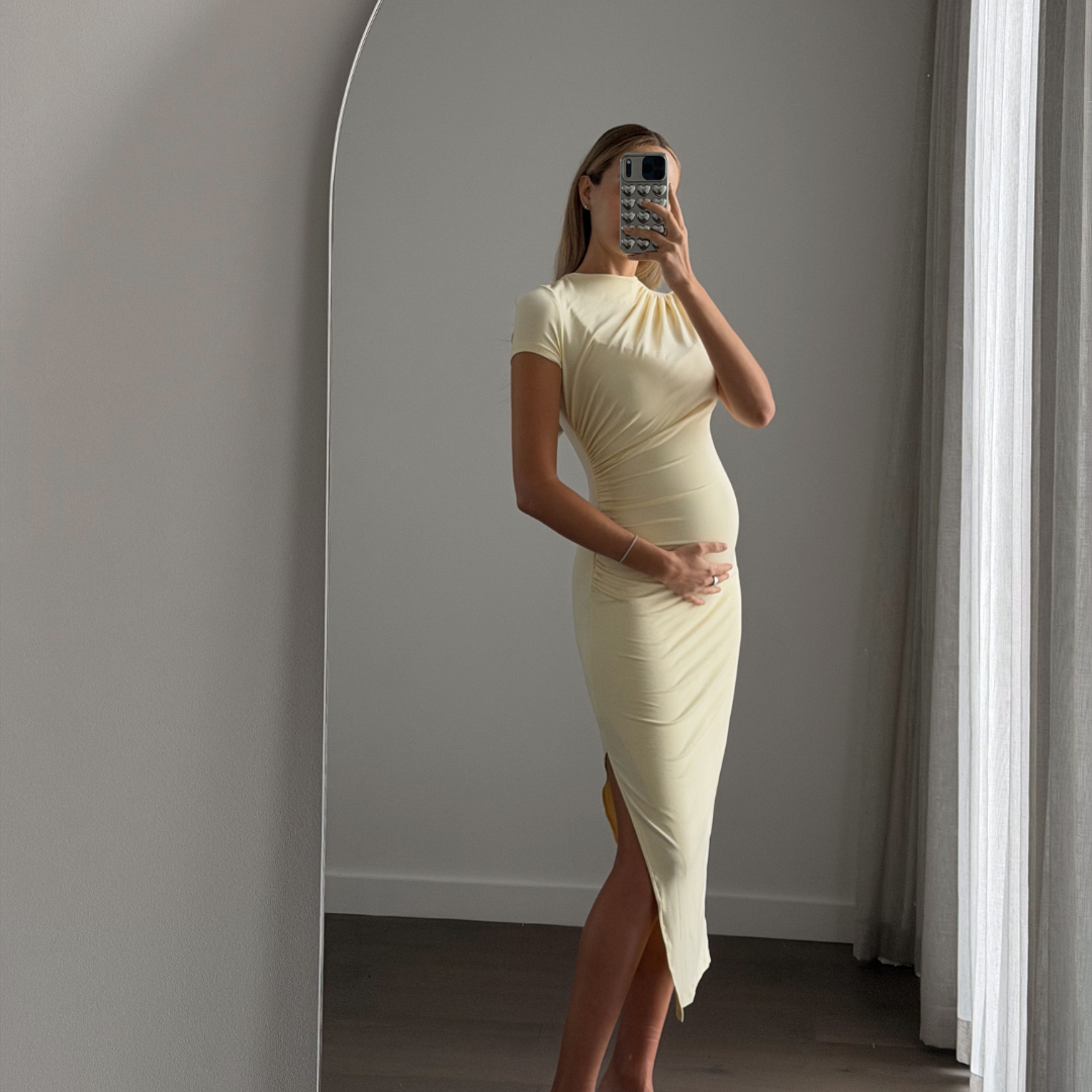 Woman wearing the Matea BEYLA baby yellow ruched midi dress showing side split, six months pregnant.